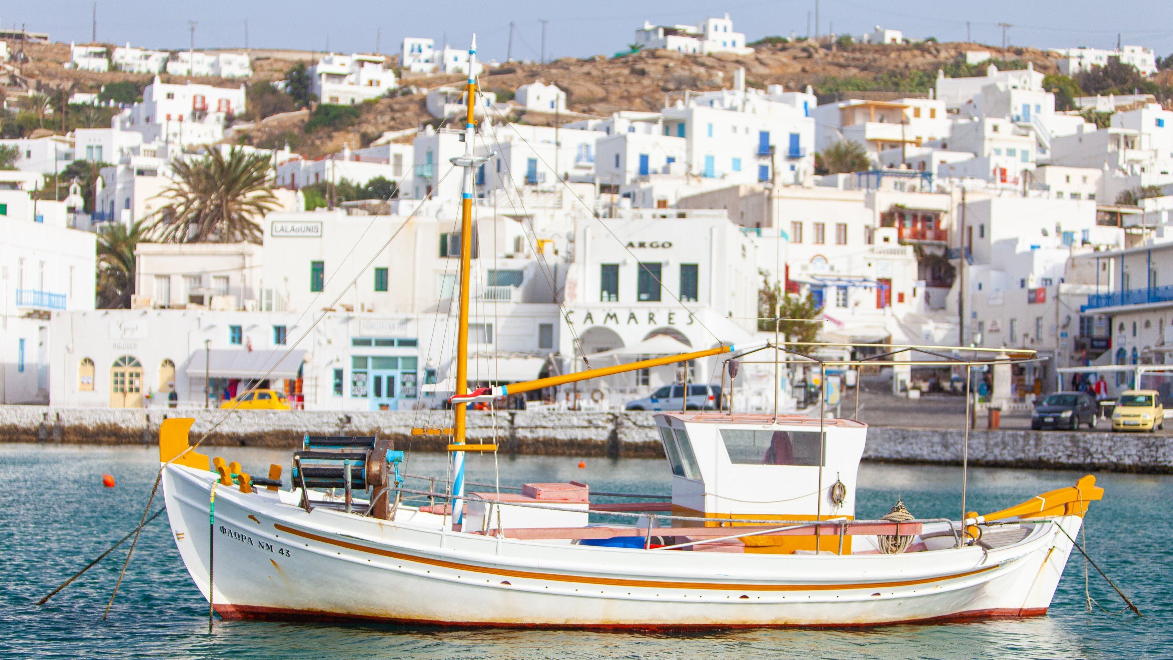 A white boat with orange accents docked near a coastal town with white buildings and a hill in the background.