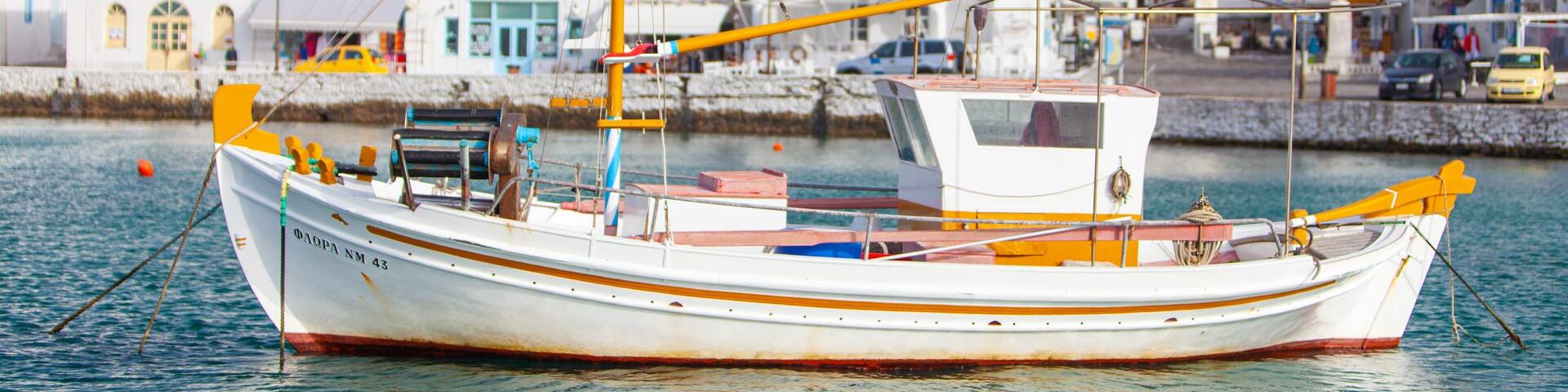 A white boat with orange accents docked near a coastal town with white buildings and a hill in the background.