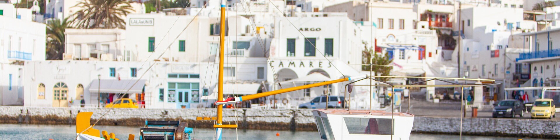 Un barco blanco con detalles naranja atracado cerca de un pueblo costero con edificios blancos y una colina al fondo.