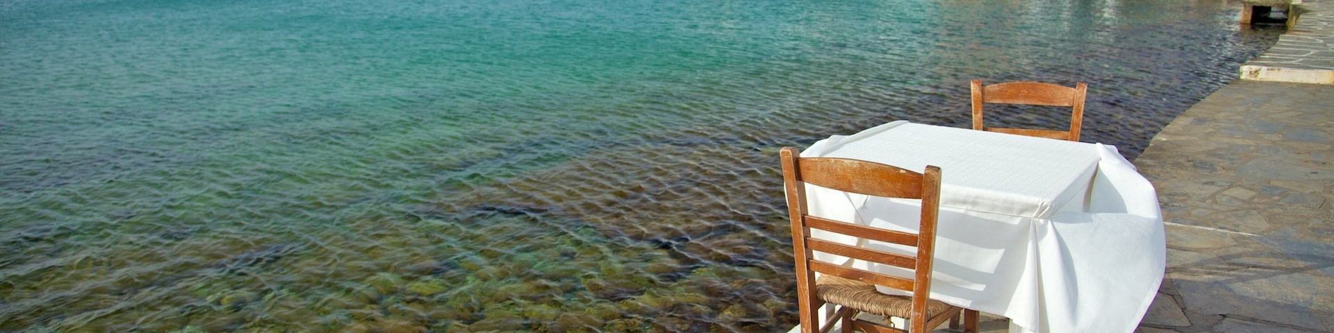 A seaside table and chairs on a stone walkway with clear water and colorful buildings in the background.