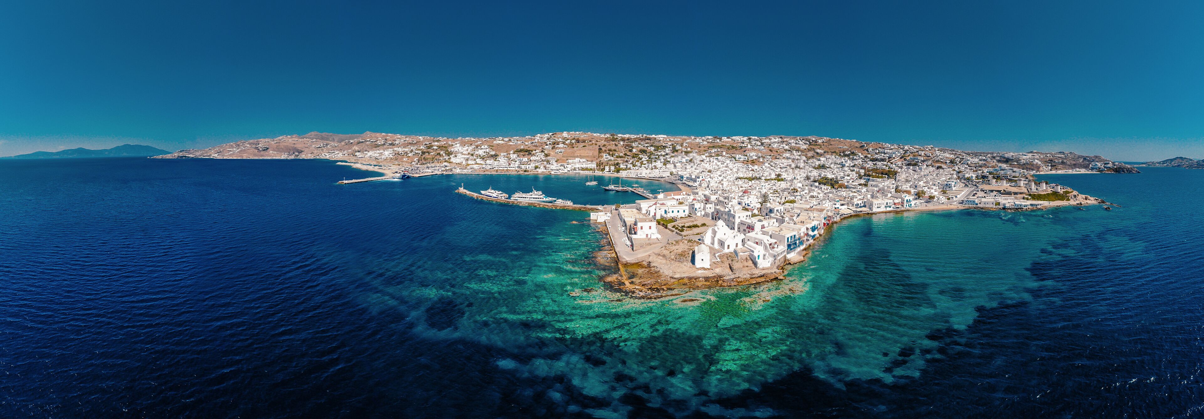 An aerial view of a coastal town with clear blue waters, white buildings, and a hilly landscape in the background.