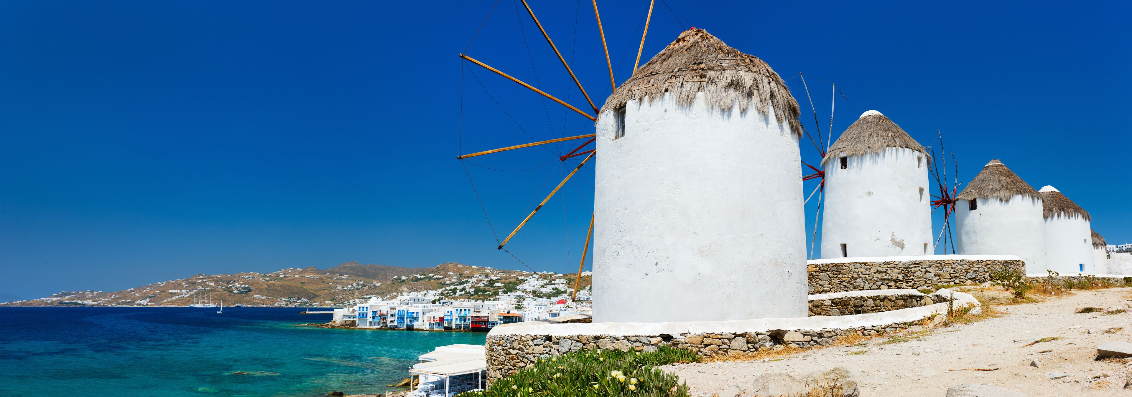 Molinos con paredes blancas y techos de paja, cielo azul despejado y un pueblo costero con edificios blancos.