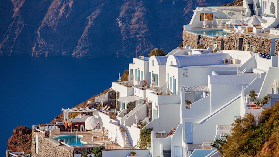 A white-washed building with a pool and outdoor seating, overlooking a steep cliff and the sea.