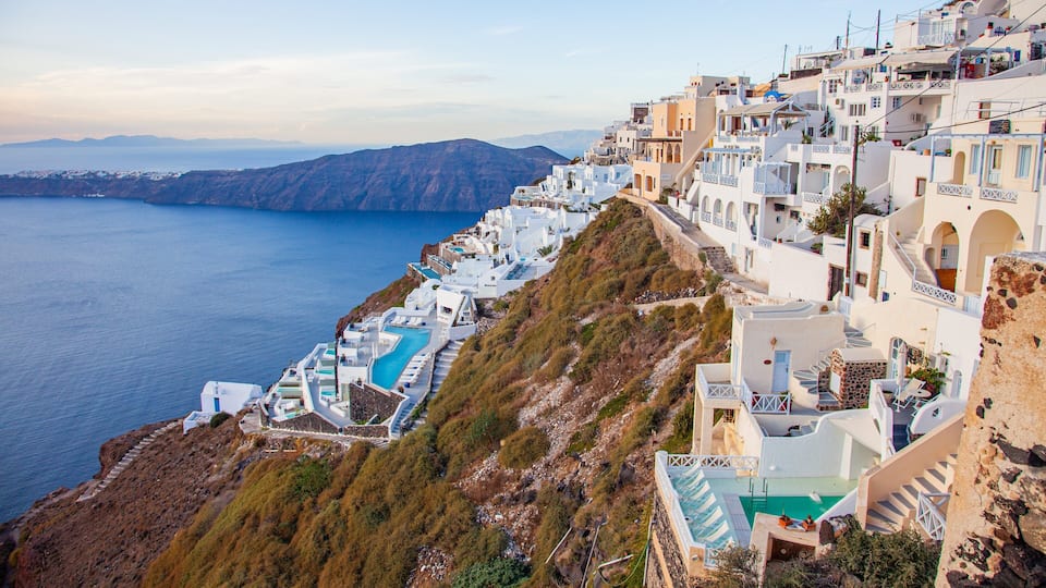 A coastal village with white buildings, a winding pool, and a view of the sea and mountains.
