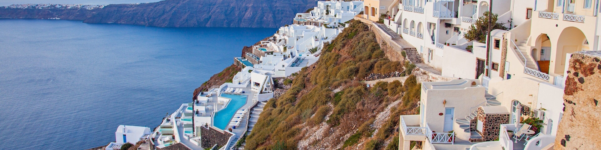 A coastal village with white buildings, a winding pool, and a view of the sea and mountains.
