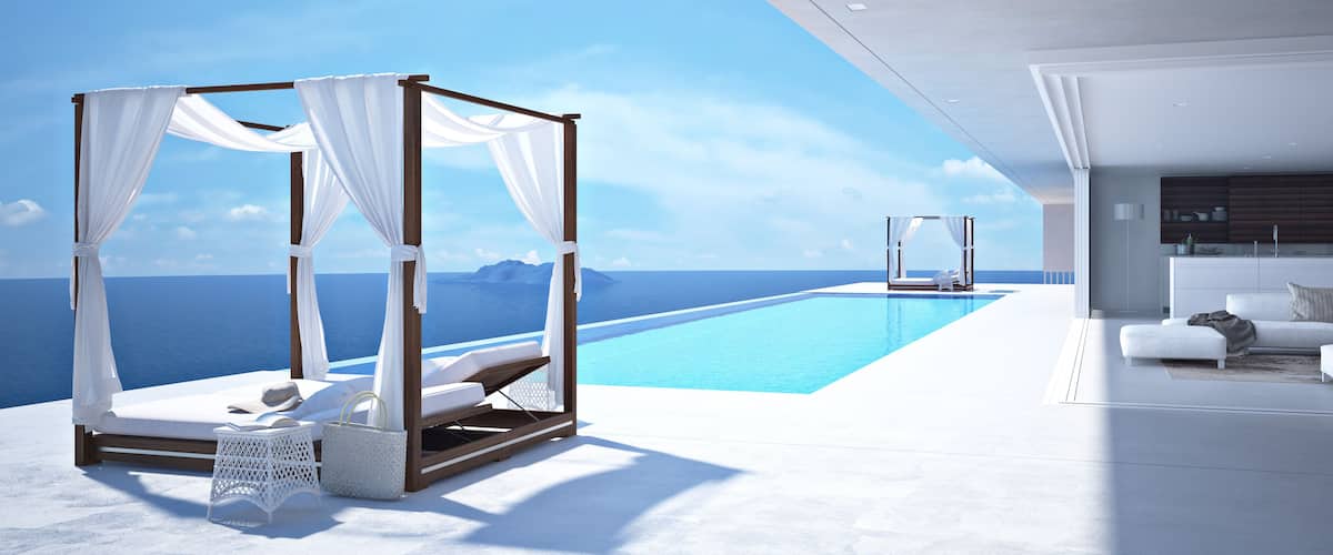 A luxurious infinity pool with a white infinity-edge deck, a modern living area with a sofa and a kitchen, and a canopy bed overlooking the ocean.