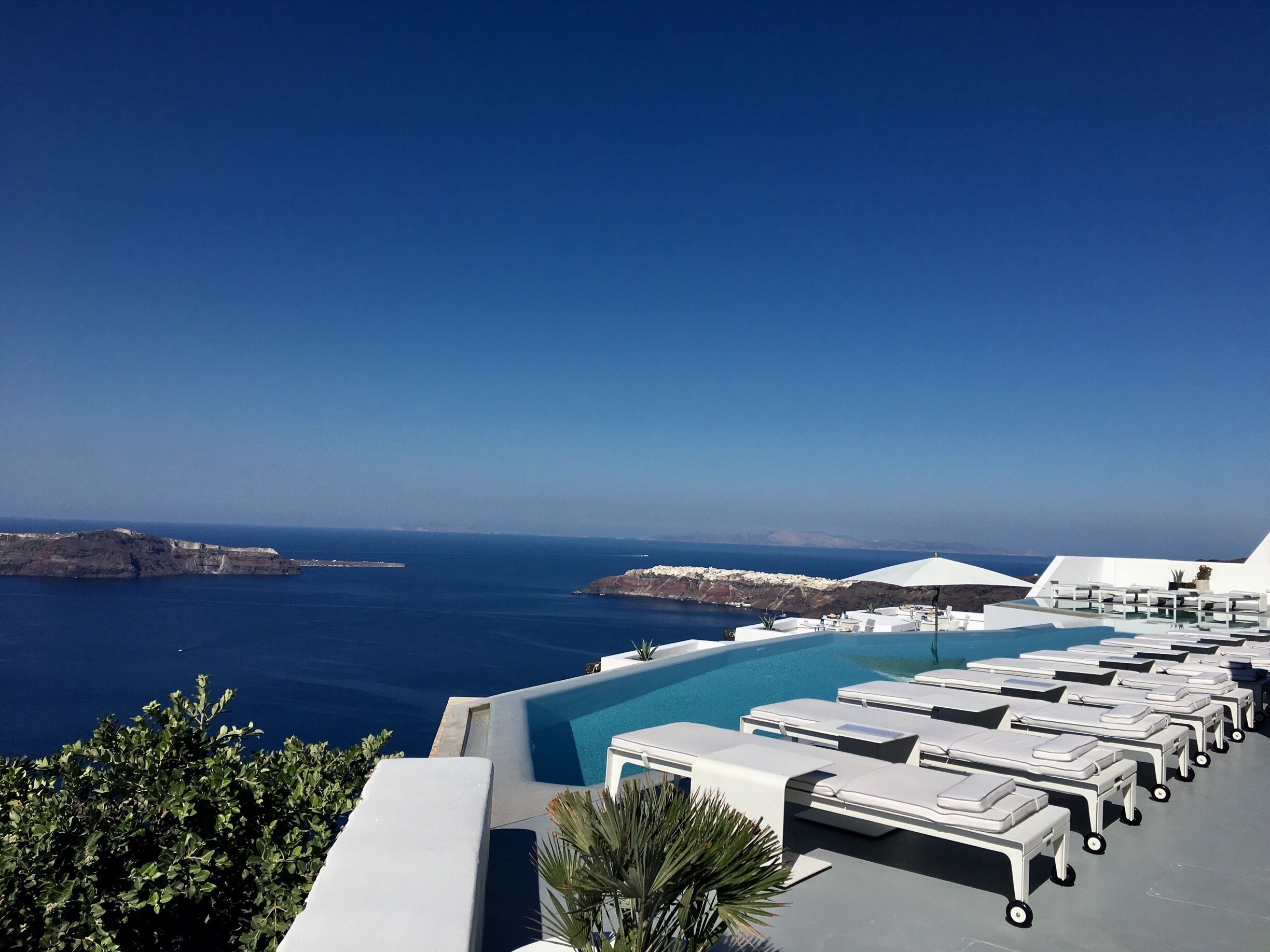 A poolside area with lounge chairs and umbrellas overlooking a clear blue sea and distant islands.