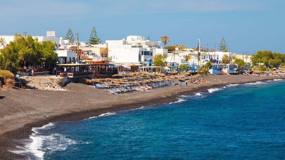 A coastal town with a beach, blue umbrellas, and white buildings.