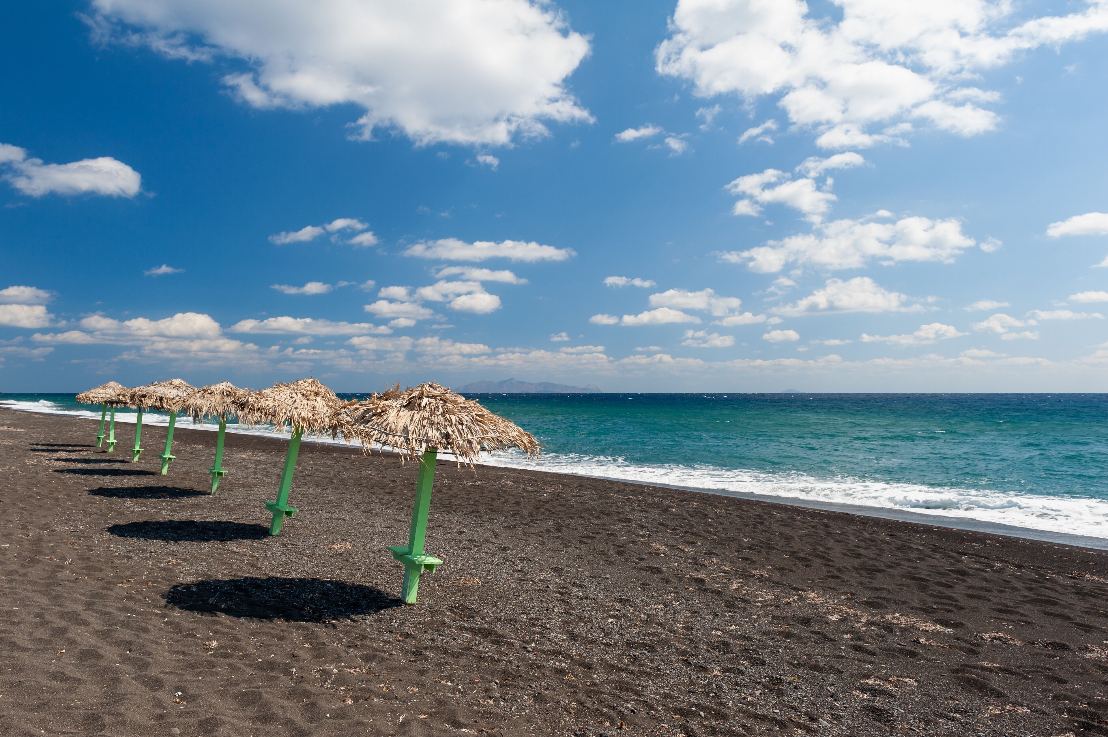 A beach with black sand and thatched umbrellas.