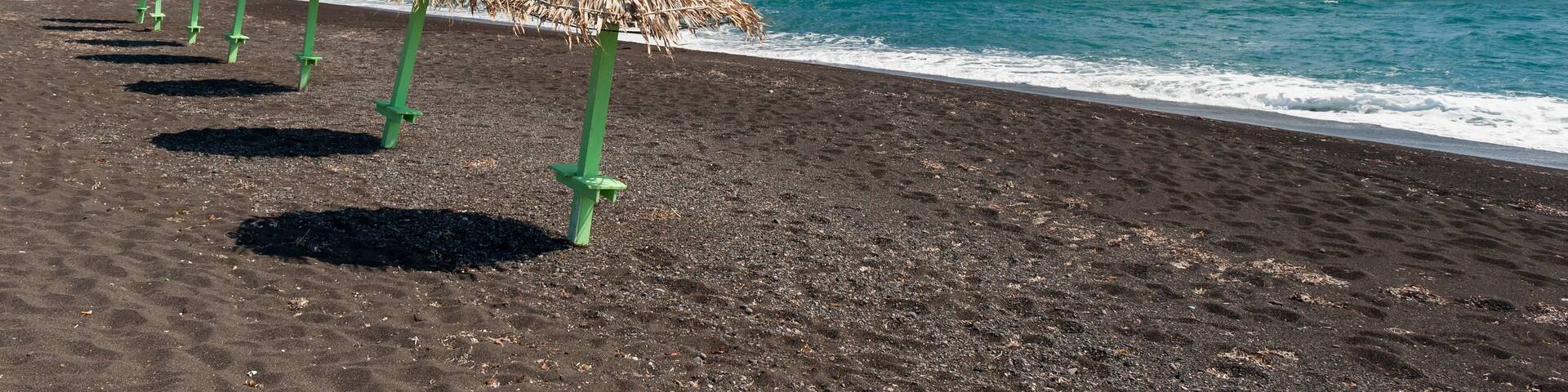 A beach with black sand and thatched umbrellas.