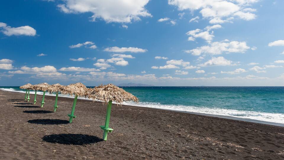 A beach with black sand and thatched umbrellas.
