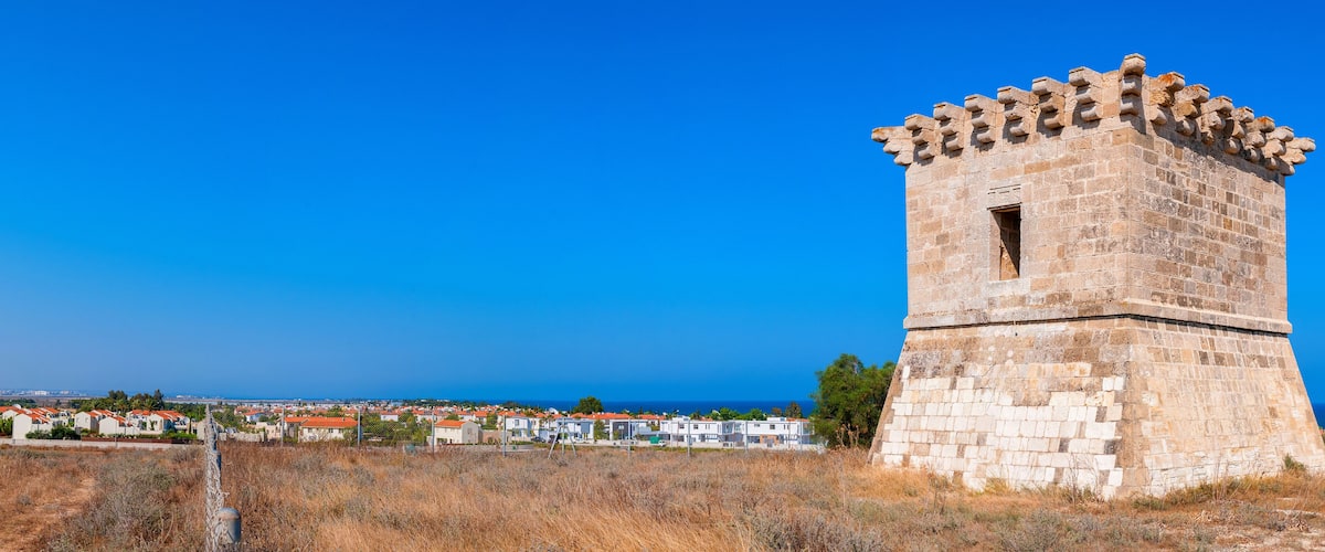 A stone tower with a crenellated top, surrounded by dry grass and a coastal town with red-roofed houses.