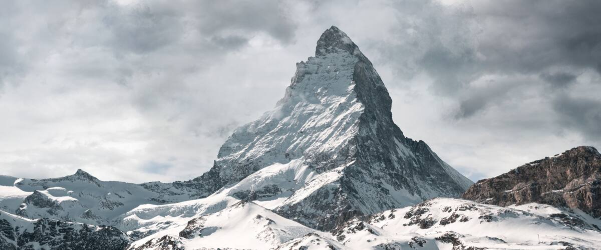 A snow-covered mountain peak with rugged terrain and a cloudy sky.