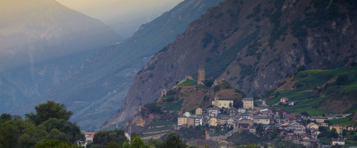 A mountainous village with a prominent tower, terraced vineyards, and a valley.