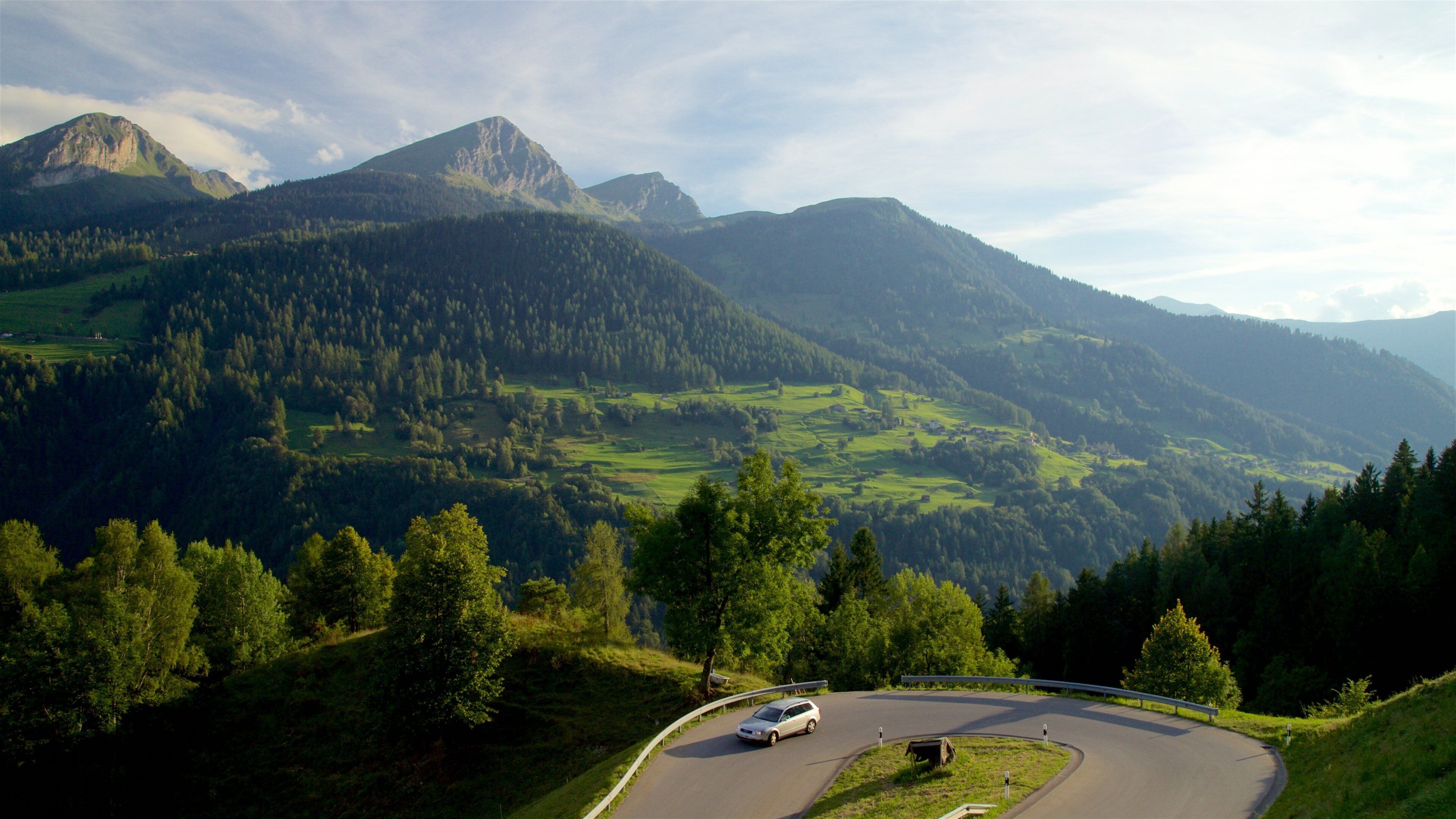 A winding mountain road with a car, surrounded by lush green hills and a prominent mountain peak in the background.