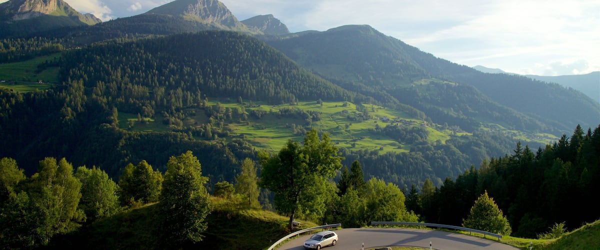 A winding mountain road with a car, surrounded by lush green hills and a prominent mountain peak in the background.