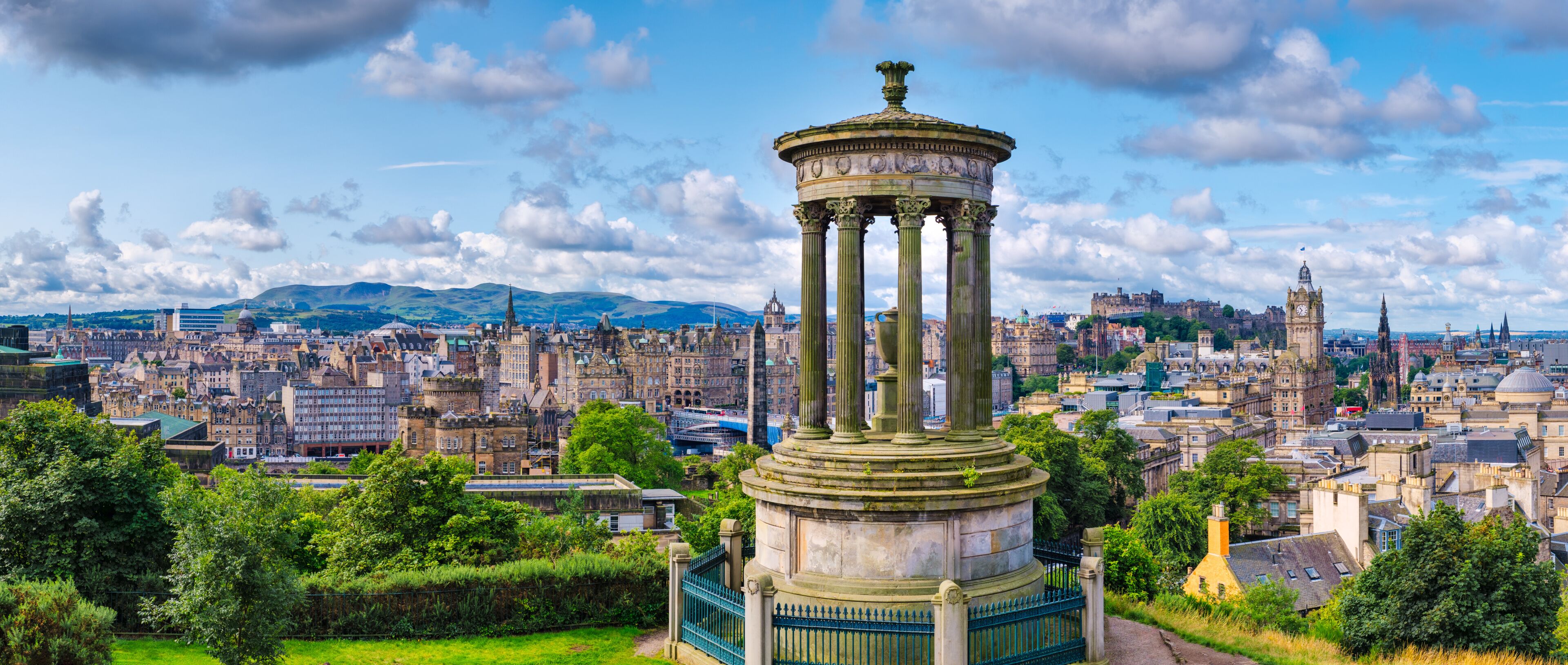 A historic monument with columns, surrounded by a cityscape with varied architecture and a distant mountain range.