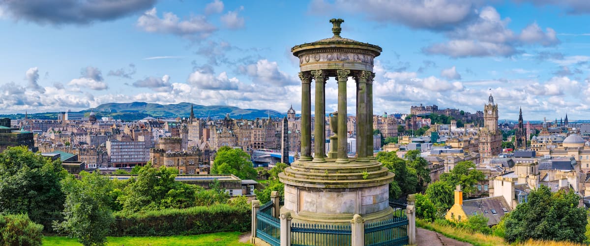 A historic monument with columns, surrounded by a cityscape with varied architecture and a distant mountain range.