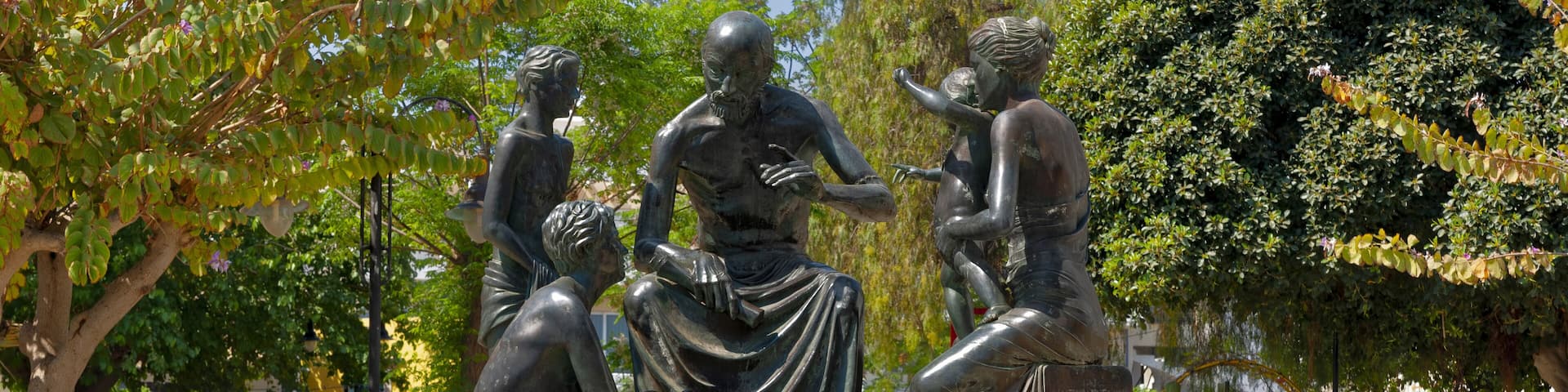 A bronze statue of a seated man with a child, surrounded by greenery and a stone wall.
