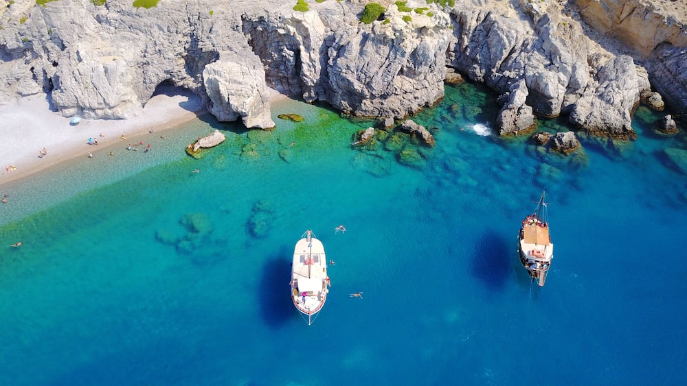 Vue aérienne d’une zone côtière avec des eaux turquoise limpides, des falaises rocheuses et deux bateaux ancrés.