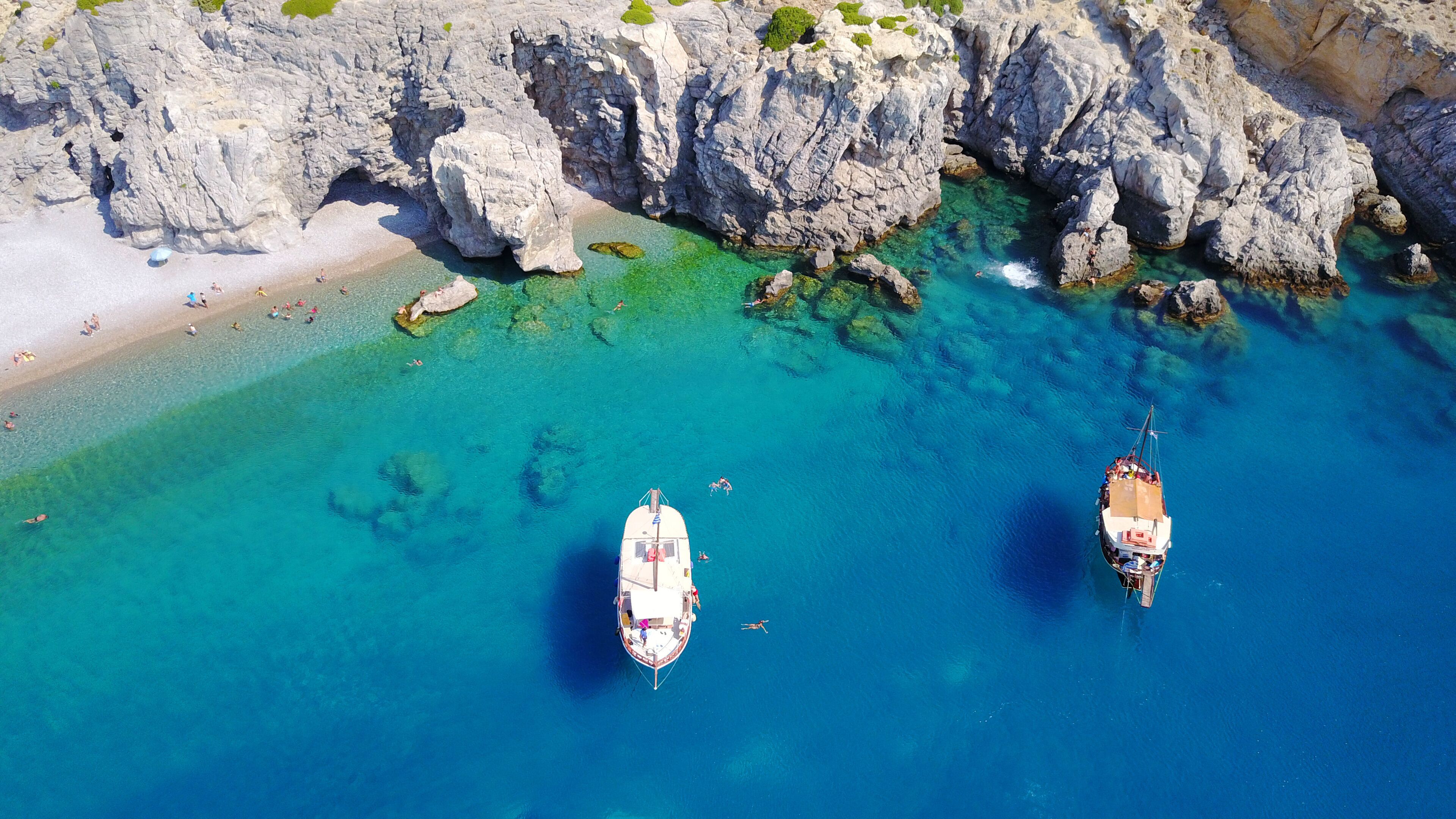 An aerial view of a coastal area with clear turquoise waters, rocky cliffs, and two anchored boats.