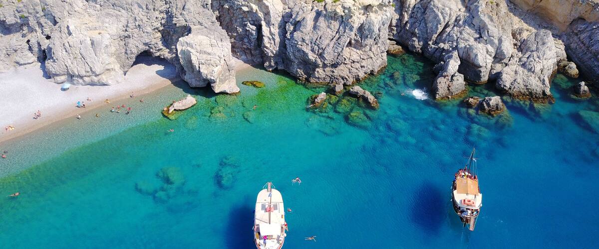 An aerial view of a coastal area with clear turquoise waters, rocky cliffs, and two anchored boats.