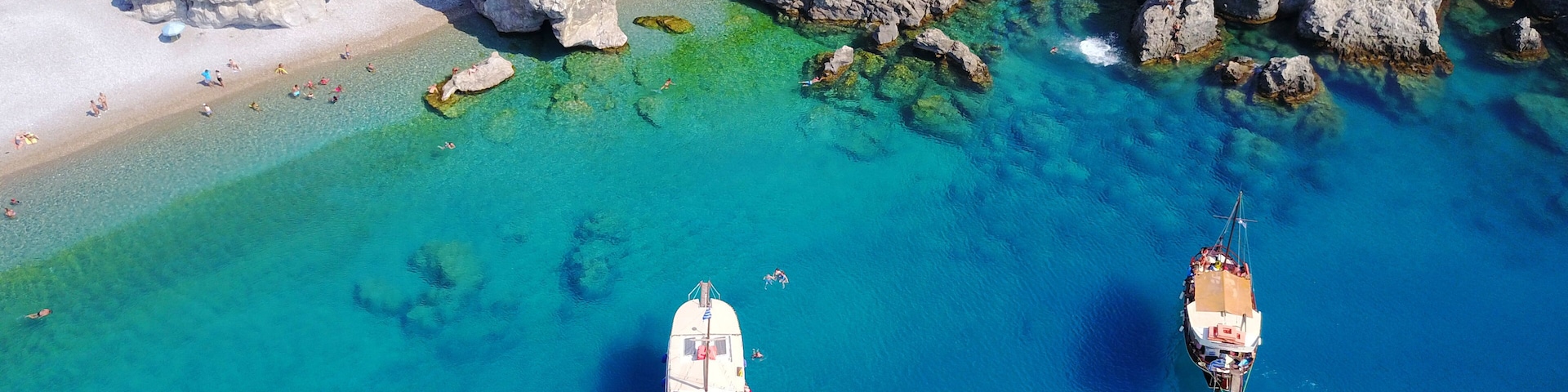 An aerial view of a coastal area with clear turquoise waters, rocky cliffs, and two anchored boats.
