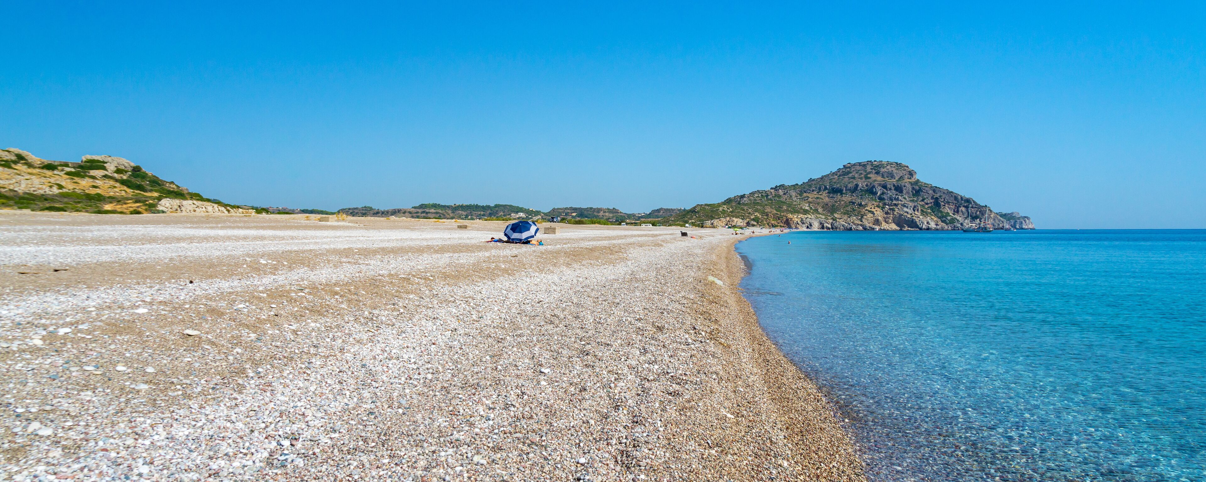 A pebbly beach with clear blue water, a distant rocky hill, and a clear sky.