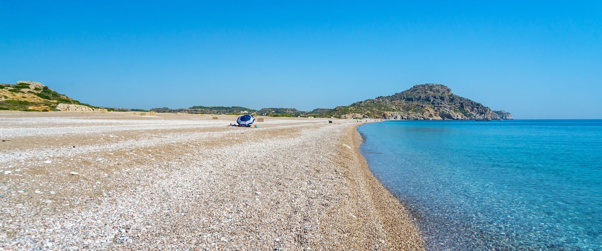 A pebbly beach with clear blue water, a distant rocky hill, and a clear sky.