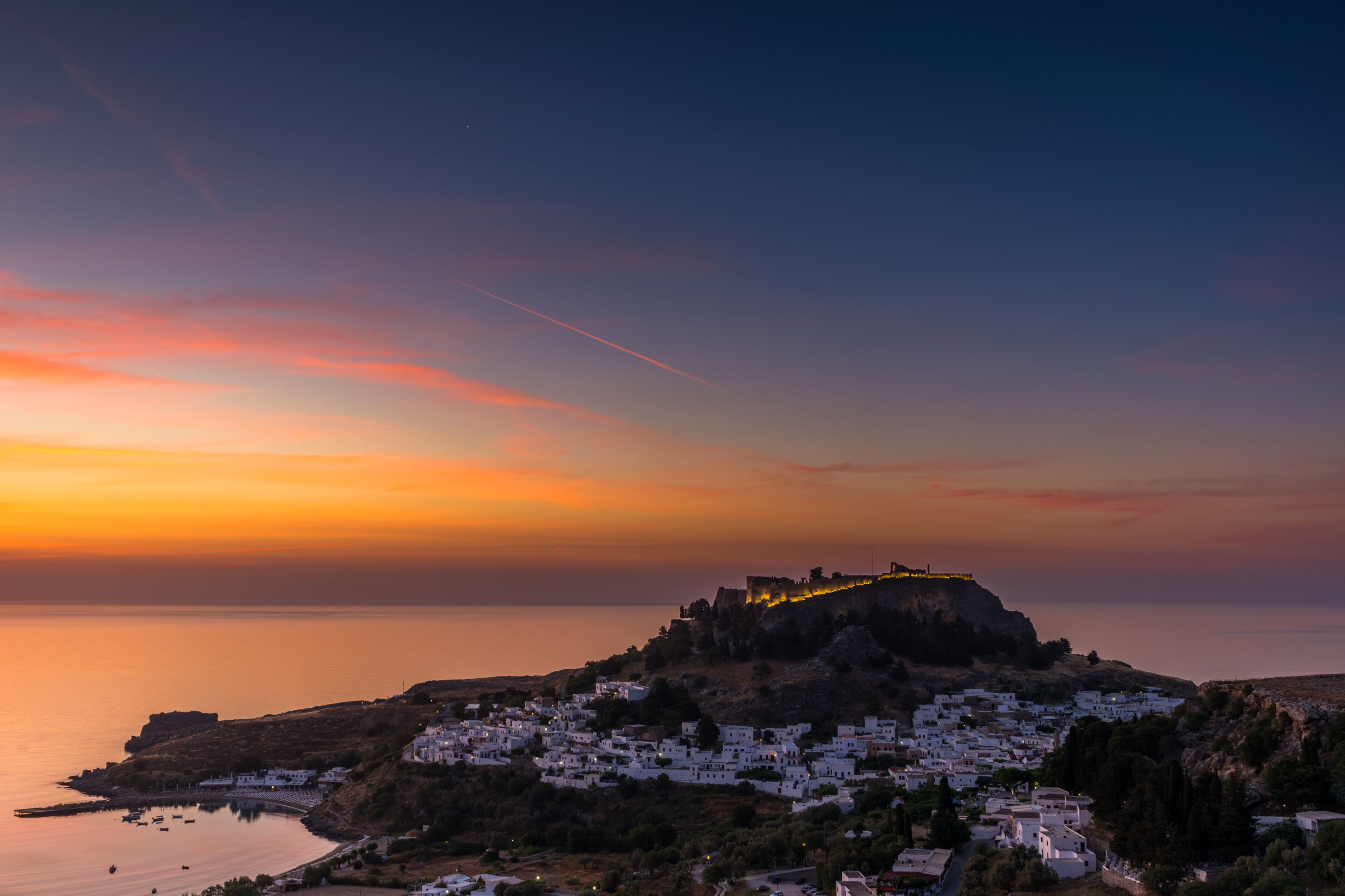 A coastal town at sunset with a prominent hilltop fortress, calm sea, and white buildings.