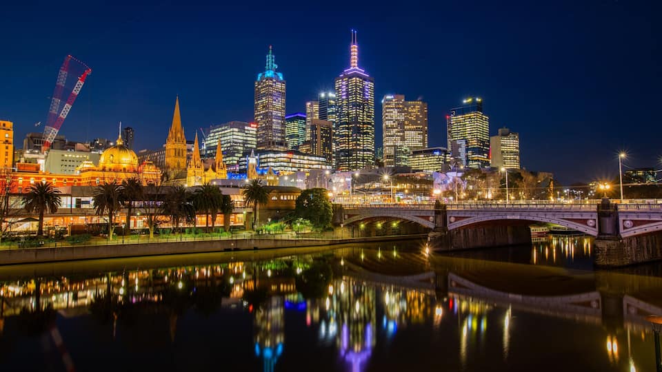 A city skyline at night with illuminated buildings, a bridge, and a river reflecting the lights.