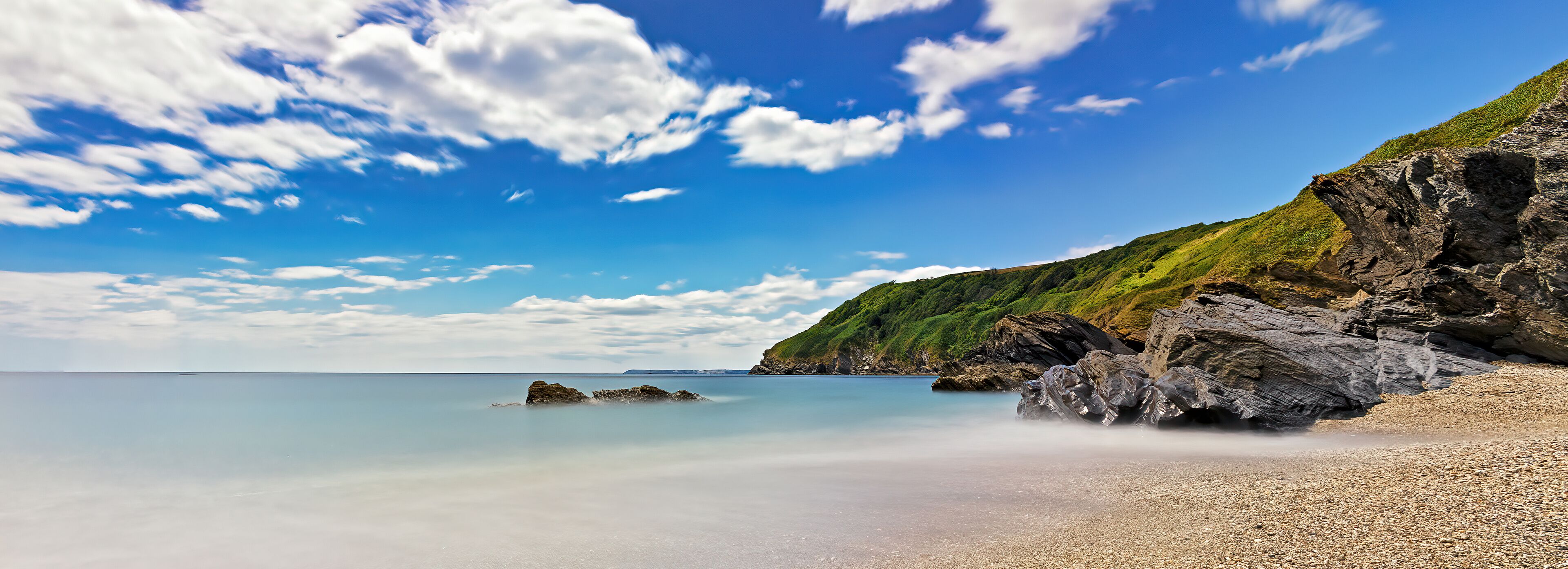 A coastal landscape with a pebbly beach, calm sea, and a rocky cliff with greenery.