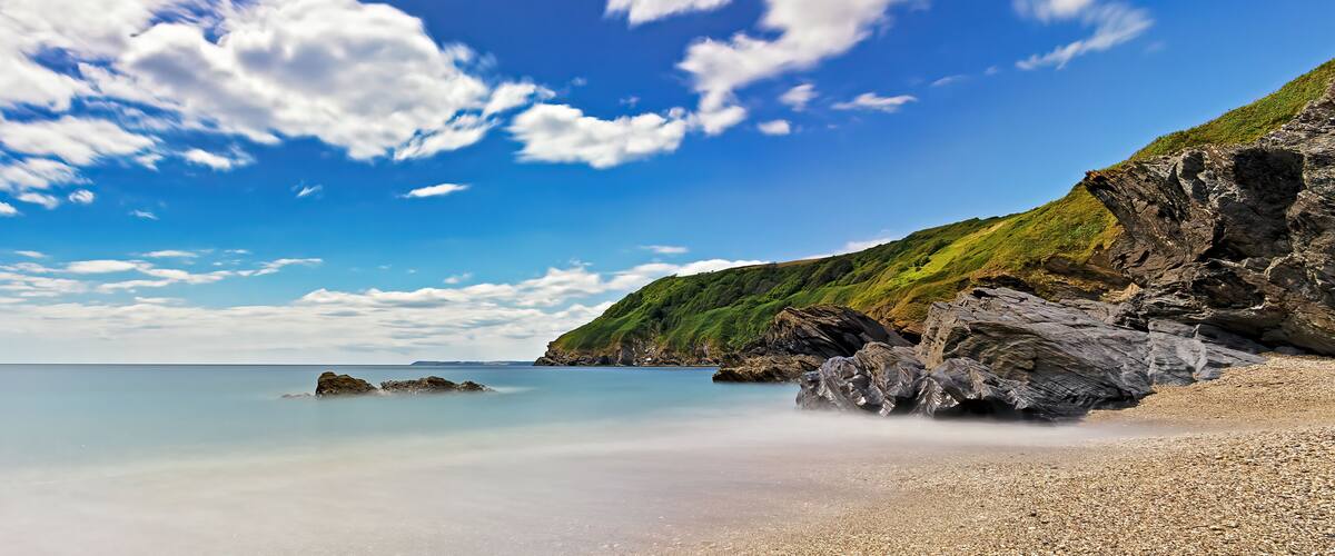 A coastal landscape with a pebbly beach, calm sea, and a rocky cliff with greenery.