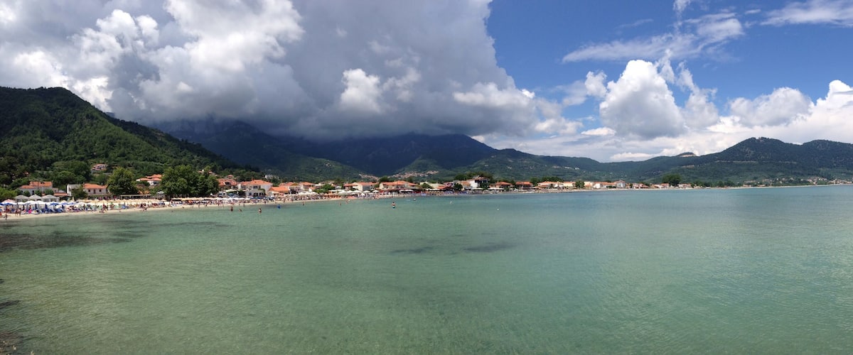 A coastal town with a clear blue sea, mountains in the background, and a rocky shoreline.
