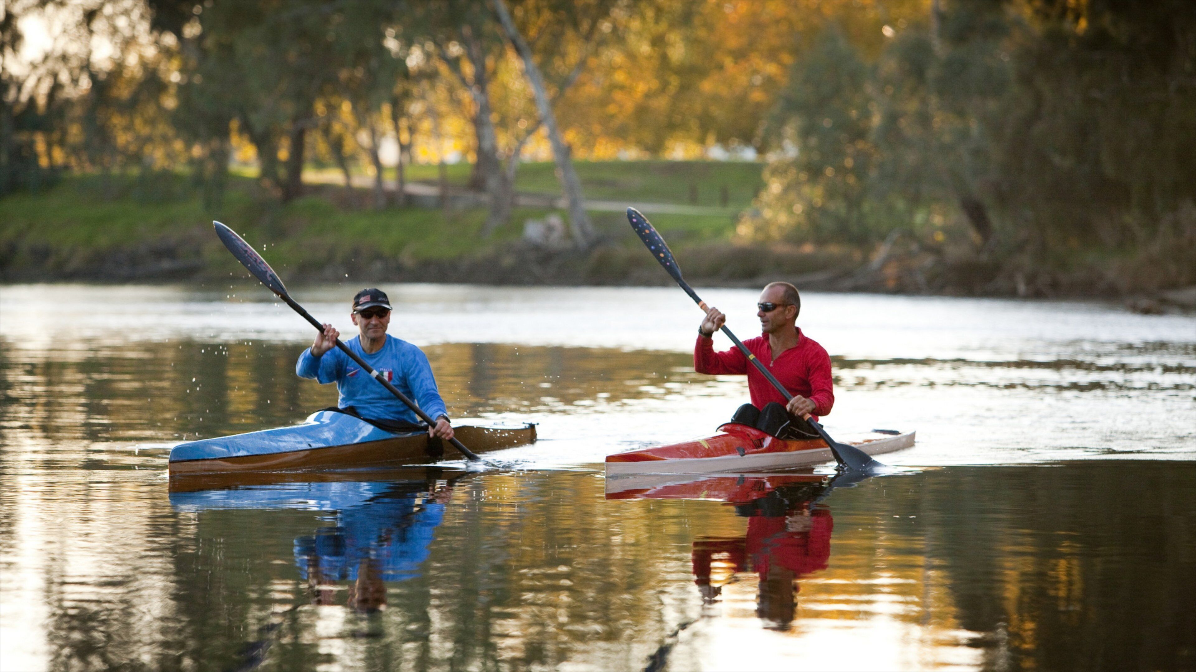 Two people kayaking on a calm river with trees in the background.