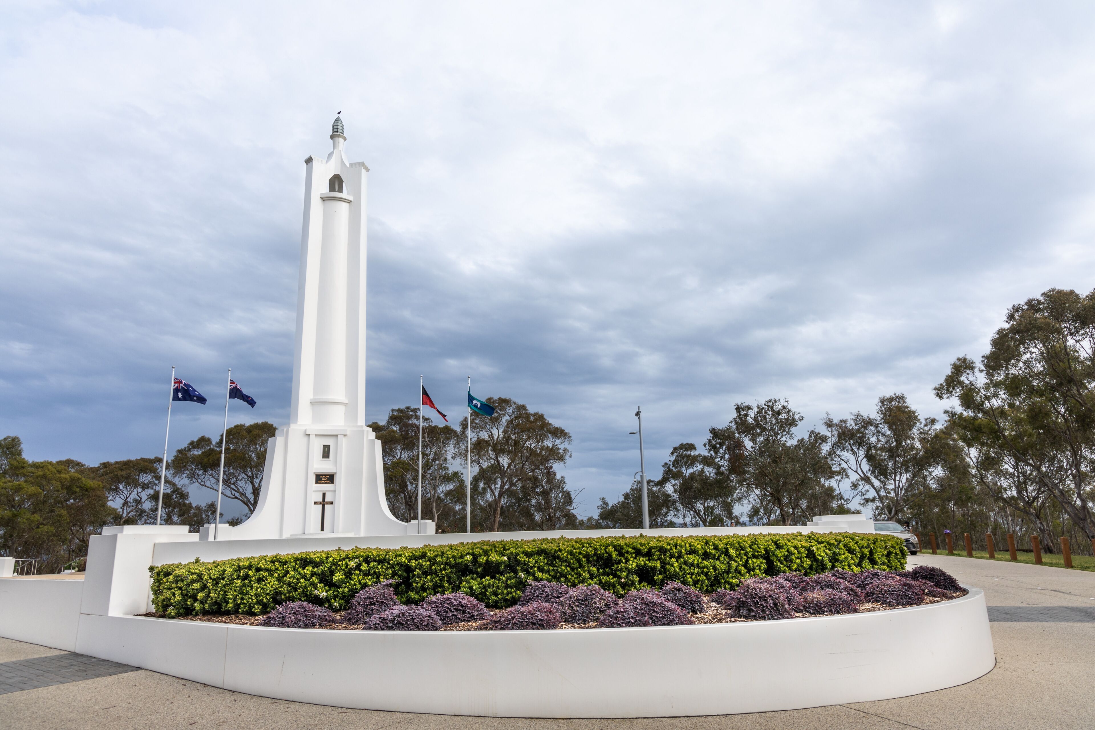 A white monument with a cross, surrounded by a circular garden with purple and green plants, under a cloudy sky.