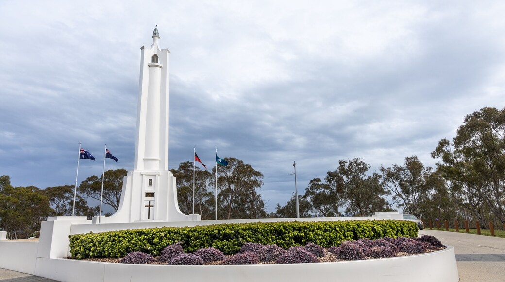 A white monument with a cross, surrounded by a circular garden with purple and green plants, under a cloudy sky.