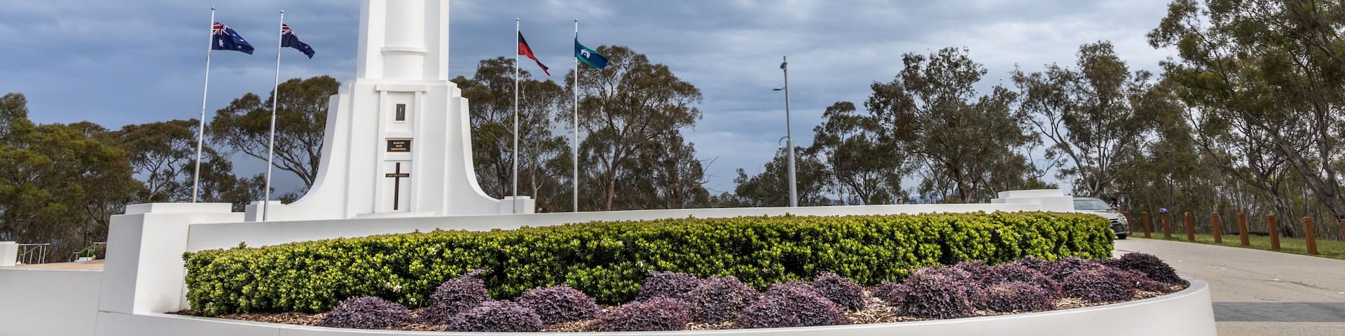 A white monument with a cross, surrounded by a circular garden with purple and green plants, under a cloudy sky.