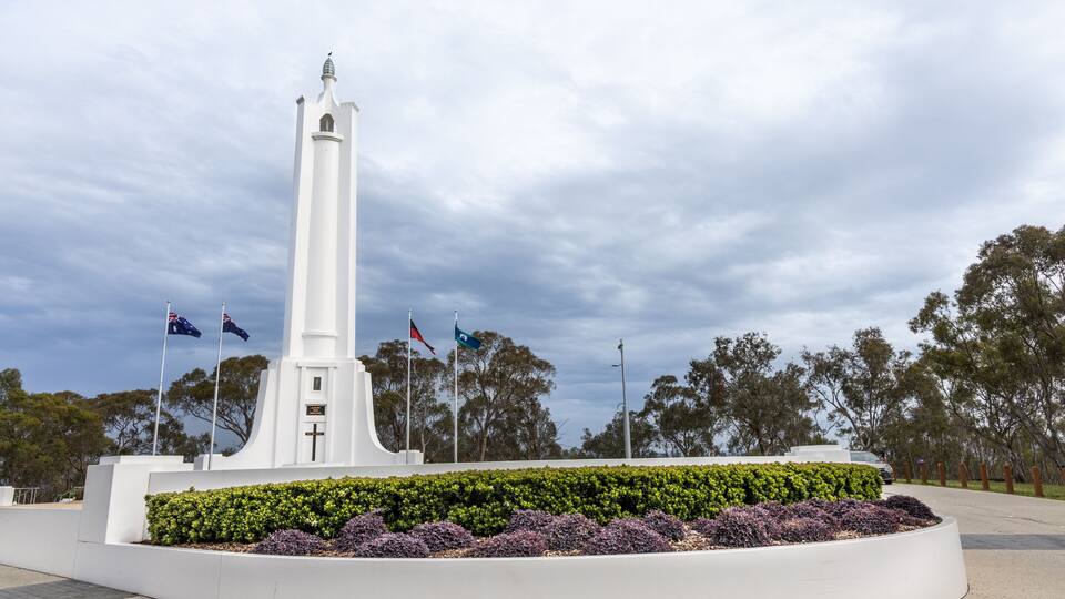 A white monument with a cross, surrounded by a circular garden with purple and green plants, under a cloudy sky.