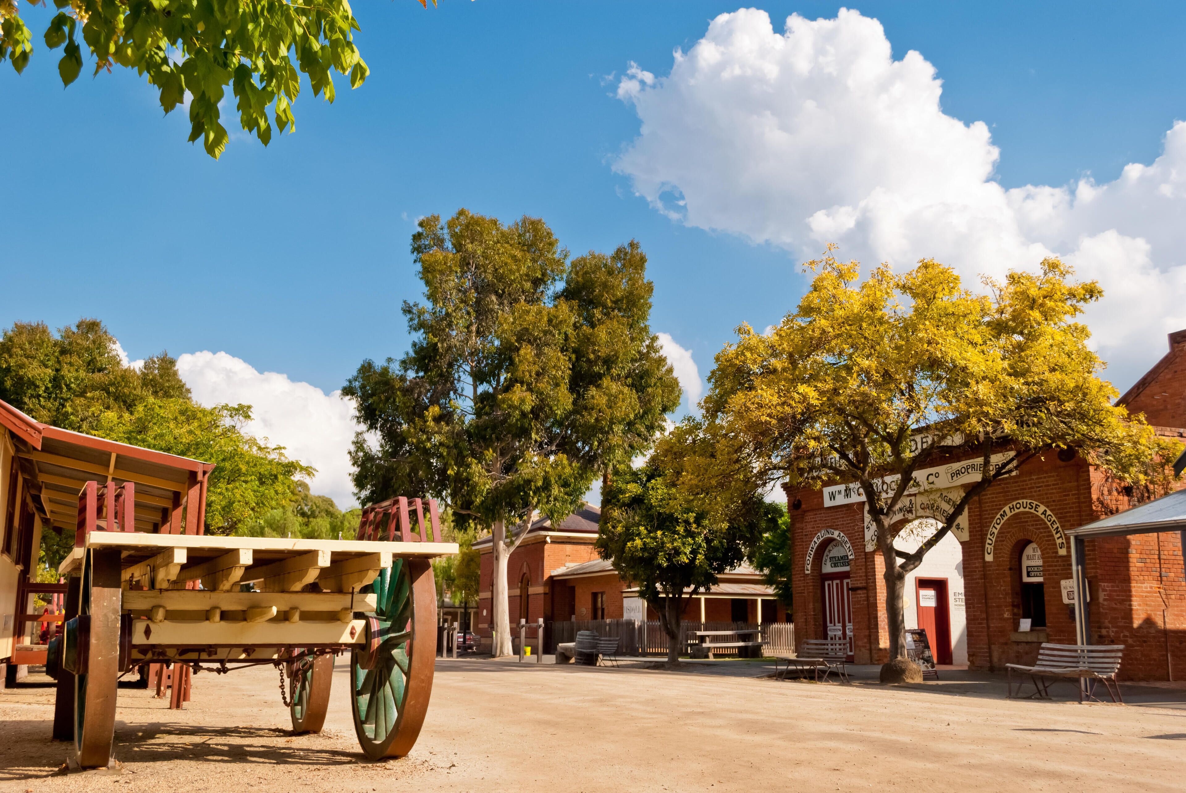 A historical town square with a wooden cart, brick buildings, and trees.