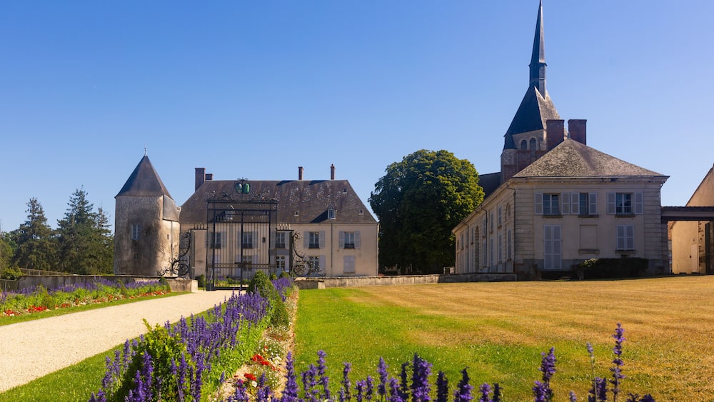 Un bâtiment historique doté d’une tour, d’un grand arbre et d’une pelouse bien entretenue agrémentée de fleurs violettes.