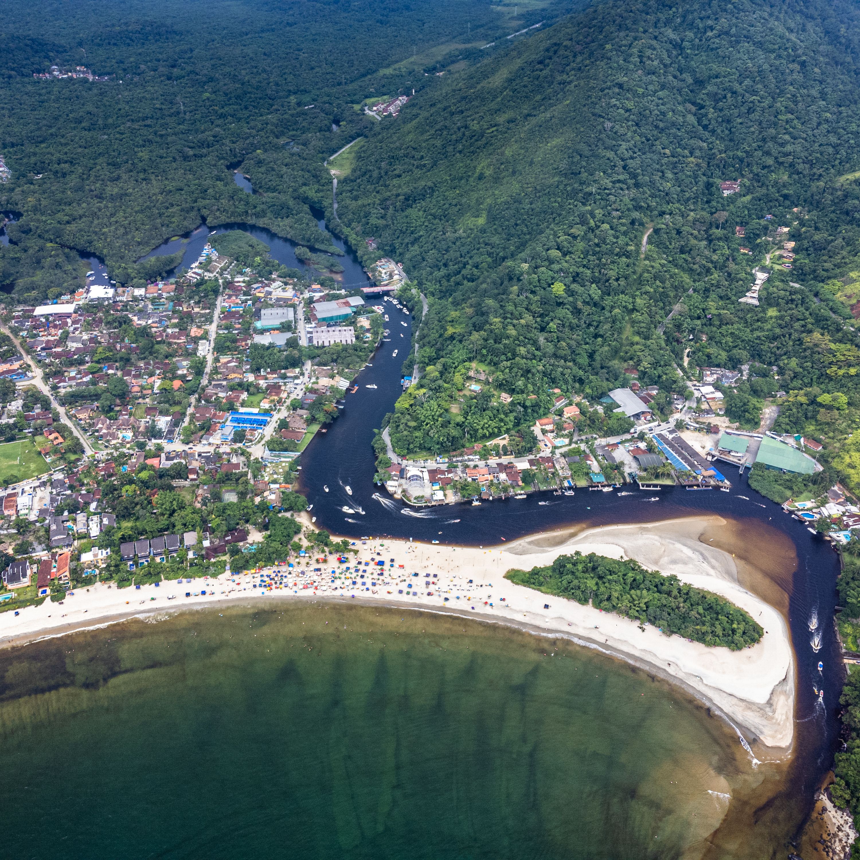 Vue aérienne d’une ville côtière avec une rivière, une forêt dense et une plage parsemée de parasols.