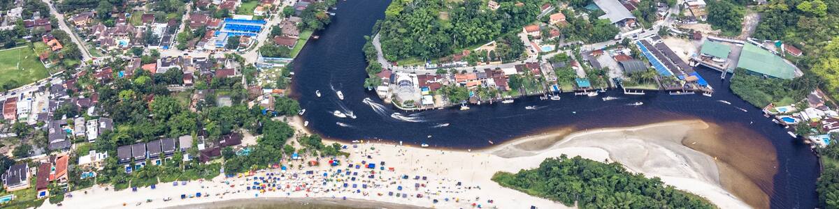 An aerial view of a coastal town with a river, dense forest, and a beach with umbrellas.
