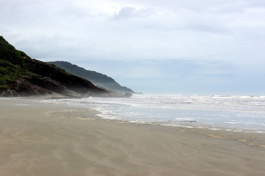 Una playa con arena, olas rompiendo en la orilla y una colina con vegetación al fondo.