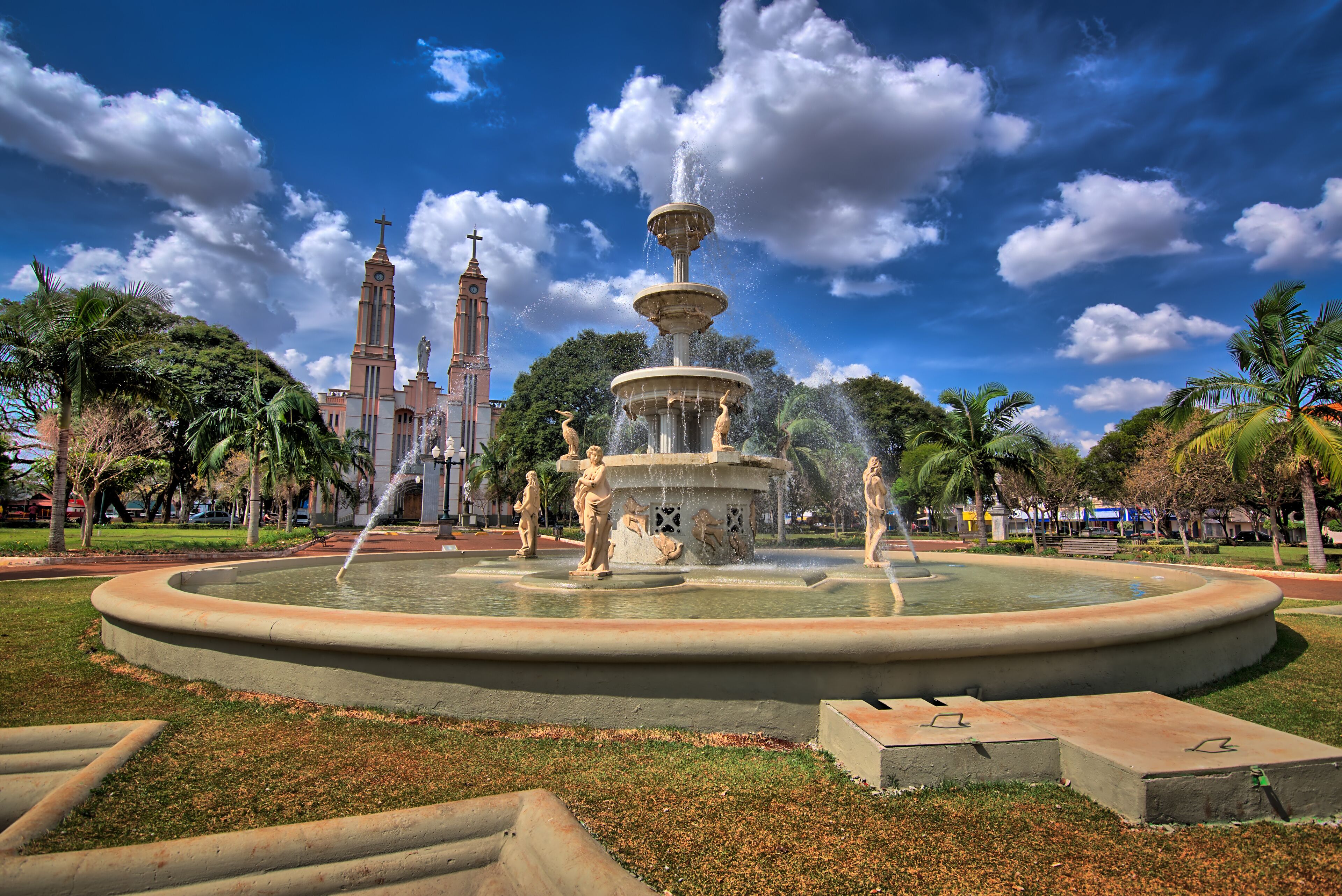 A large fountain with statues, surrounded by palm trees and a church with two towers in the background.