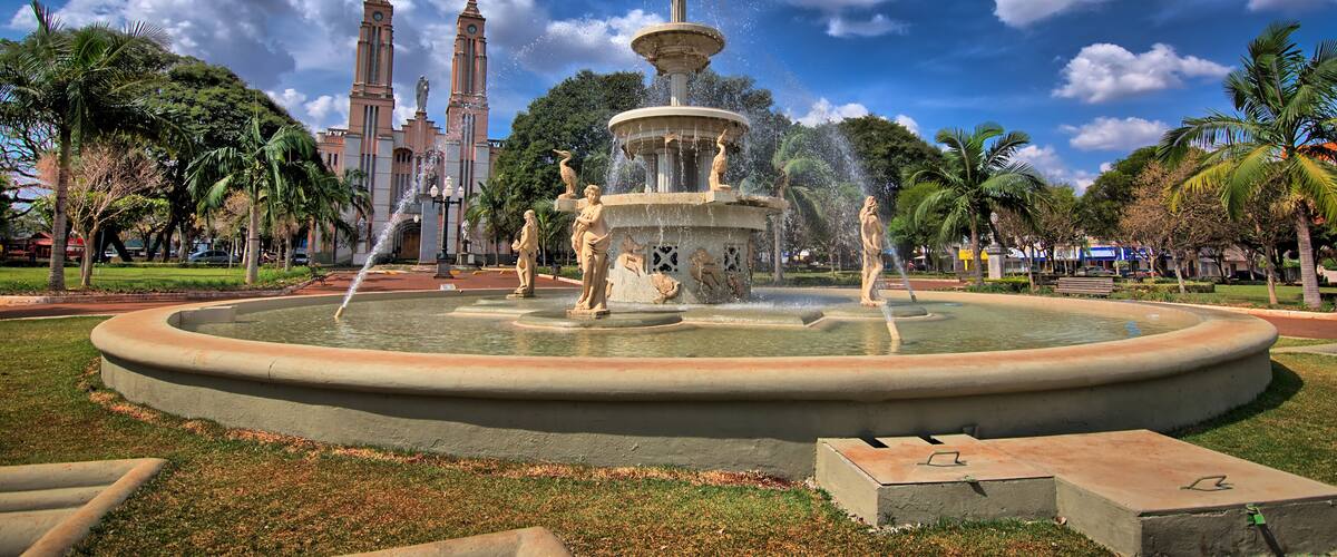 A large fountain with statues, surrounded by palm trees and a church with two towers in the background.