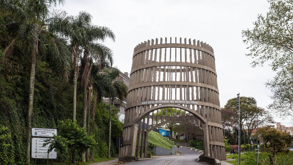 A wooden archway with a sign reading 'Bento Goncalves' and a road leading through a green area with trees.