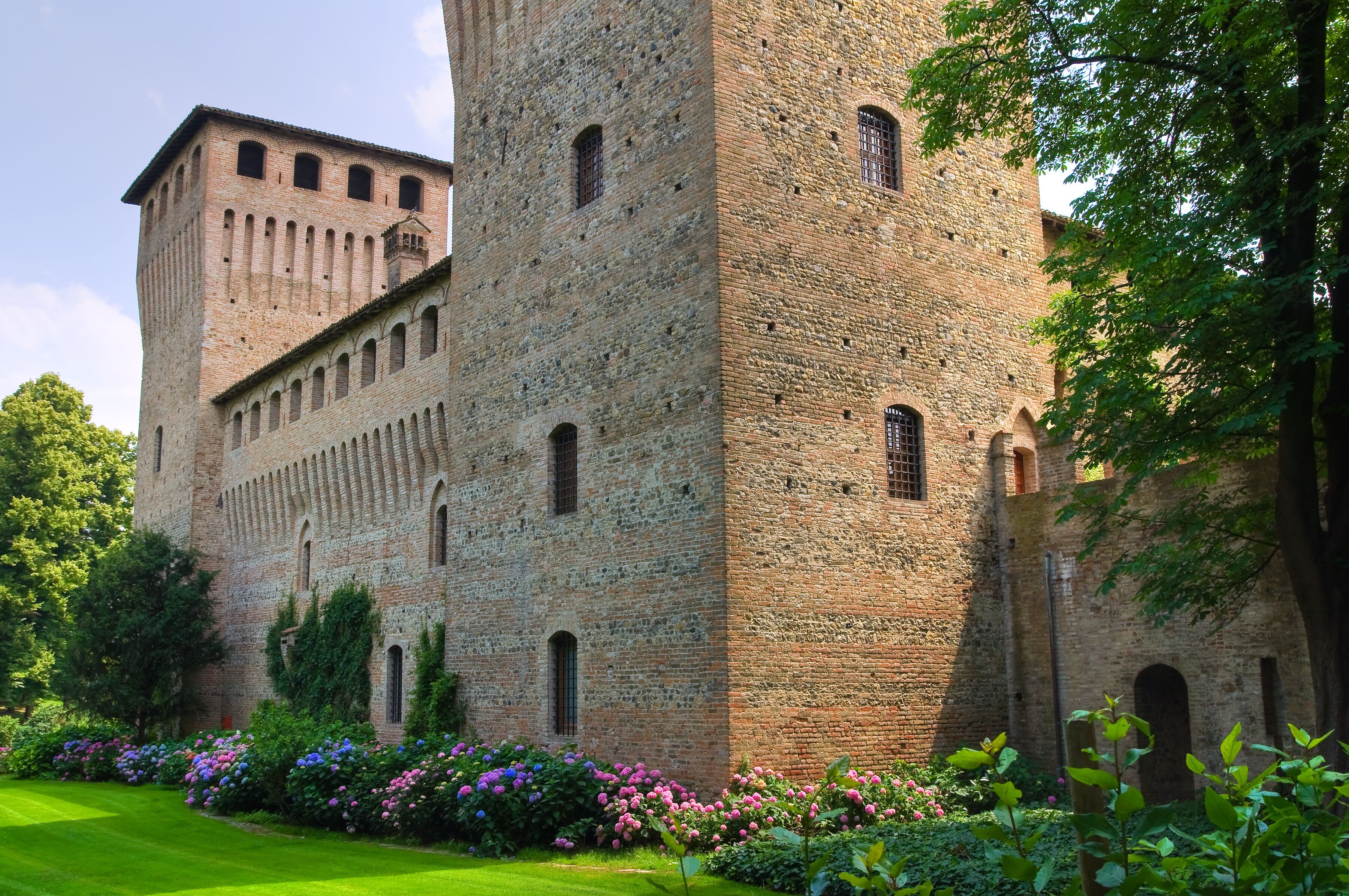 A historic brick castle with arched windows and a tower, surrounded by lush greenery and flowering plants.
