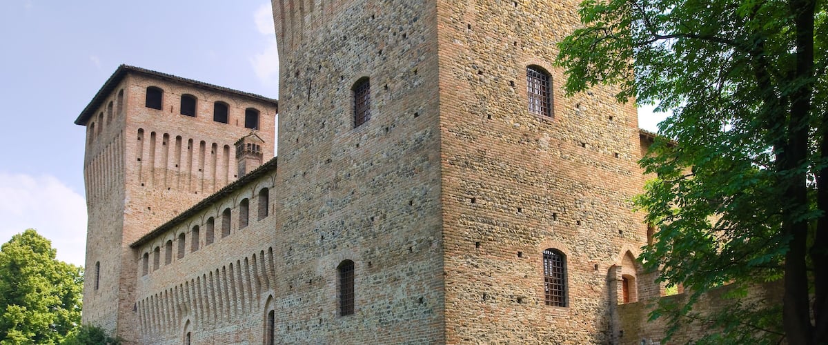 A historic brick castle with arched windows and a tower, surrounded by lush greenery and flowering plants.
