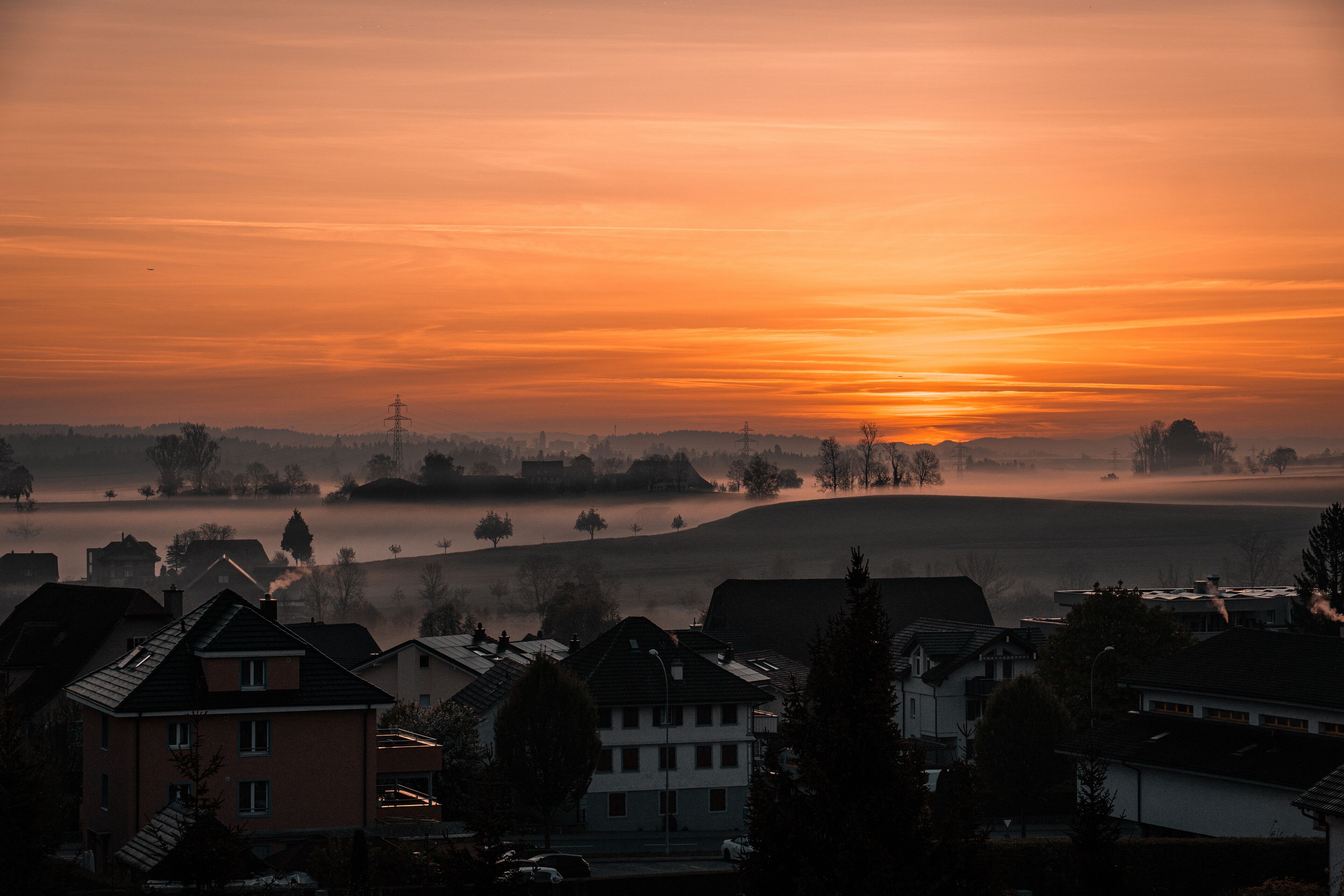 A sunrise over a misty landscape with houses and a power line tower.